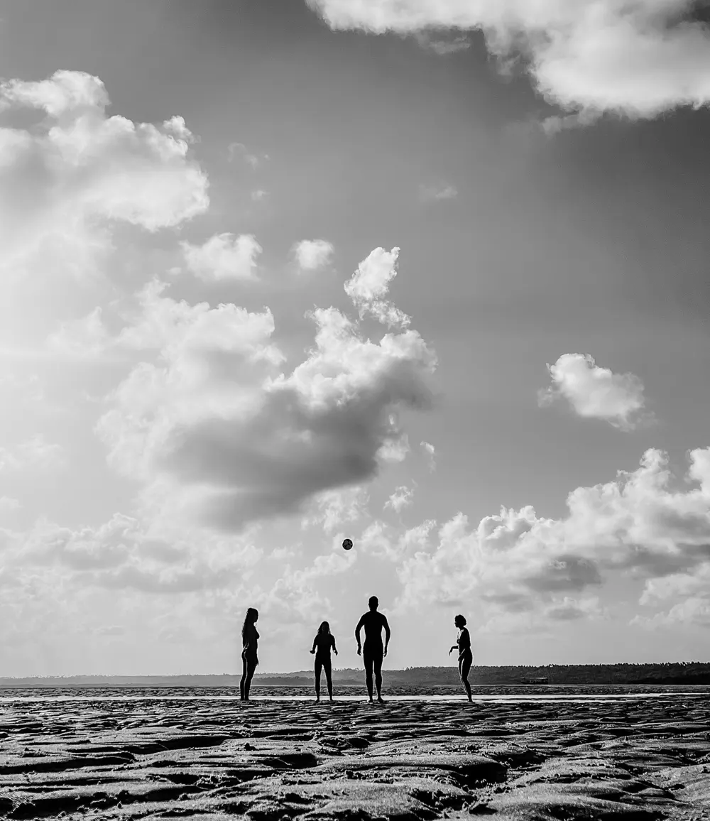 Silhuetas de quatro pessoas jogando bola na areia sob um céu de nuvens, em preto e branco
